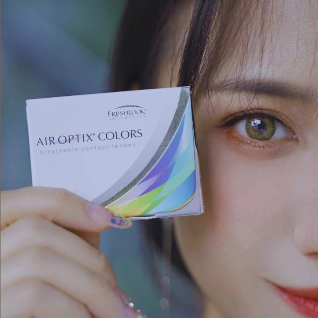 Model holding a box of Alcon Air Optix Colors contact lenses, wearing monthly brown-colored lenses