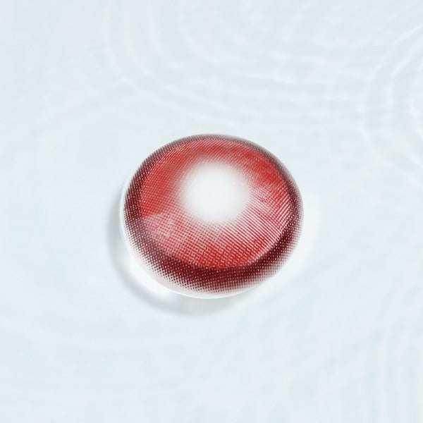 Close-up of a red colored contact lens with detailed design, resting on a reflective surface, ideal for cosplay or bold eye enhancements.